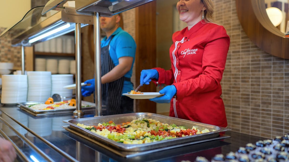 Chef in red uniform serving food at a buffet counter in Antalya restaurant, smiling and engaging with customers.